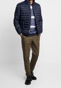 Granatowa puchowa kurtka z pionowymi przeszyciami, wysoki kołnierz, w zestawie z niebieską bluzą tie-dye i oliwkowozielonymi spodniami. Czarne buty na sznurowadła.