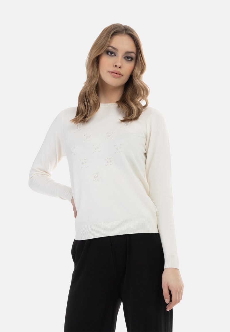 Valkoinen neulottu villapaita, jossa on pitkät hihat, teksturoitu yksityiskohta ja ribbineulehelma. Yhdistetty mustiin housuihin studio-olosuhteessa.