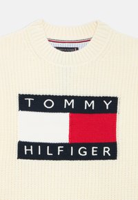 Pulover tricotat crem, cu un patch mare logo multicolor cu un design dreptunghiular în nuanțe de albastru închis, alb și roșu, amplasat în centrul pieptului.