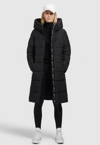 Lang zwarte pufferjas met een capuchon, voorzien van een tweewegsrits, zijzakken en contrasterende branding langs de rits, gecombineerd met zwarte leggings en sneakers.