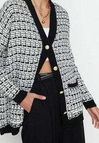 Schwarz-weiß gemusterte Strickjacke mit strukturierter Strickoptik, einem V-Ausschnitt, drei goldenen Knöpfen und Details mit einer Tasche vorne.