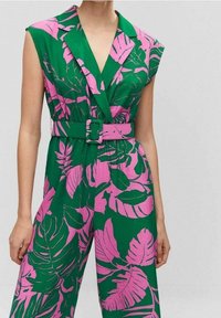 Tuta verde con un motivo di foglie tropicali rosa, dotata di un ampio colletto, vita con cintura e maniche a cappuccio; il tessuto sembra leggero e liscio.