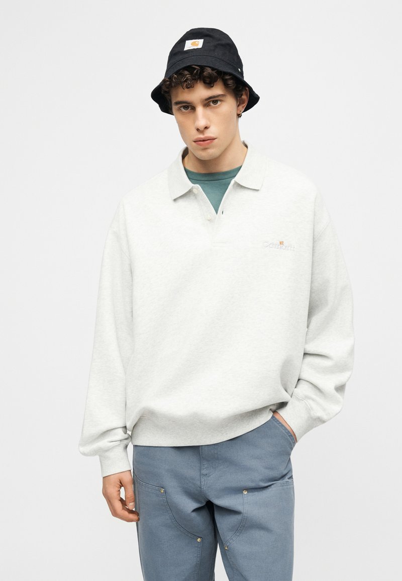 Young man wearing a white long-sleeve polo shirt, blue pants, and a black bucket hat, posing with one hand in pocket.