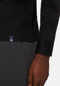 Czarna koszula z długim rękawem, z dopasowanym mankietem, z małą niebieską metką z białą literą 'B' u dołu. Gładka tekstura, dopasowany krój.