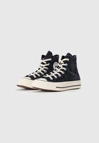 Adidași negri înalți cu un model texturat, șireturi crem, tălpi din cauciuc și o etichetă cu logo pe gleznă. Design clasic din pânză.