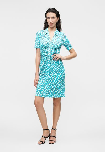 Γυναίκα που φοράει ένα μίνι τυρκουάζ φόρεμα με γεωμετρικό σχέδιο, στέκεται με το ένα χέρι στη μέση και μαύρα σανδάλια με τακούνι.