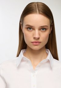 Camicia bianca con colletto, realizzata in un tessuto leggero e liscio con un leggero luccichio. Design classico con chiusura a bottoni standard.