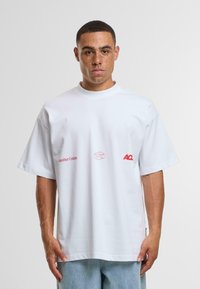 Bílý bavlněný tričko s oversized střihem, krátkými rukávy, kulatým výstřihem, a červenými potisky log na přední straně, textilní struktura a ležérní styl.