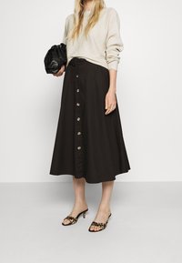Schwarzer Midi-Rock mit vorderem Knopfverschluss, kombiniert mit einem hellbeigen Pullover und offenen Mules mit Tierdruck. Schwarze Clutch-Tasche in der Hand.