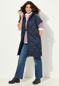 Námořnický prošívaný dlouhý vest s kapucí, který má zipové zapínání a boční patentky. Nositý přes růžový svetr a modré džíny, doplněný černými botami.