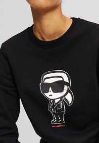Sudadera negra con una figura de dibujos animados que lleva gafas de sol, en acentos blancos y rojos, impresa en el frente. Tejido suave con cuello redondo.