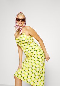 Vestido floral amarelo com uma silhueta justa, apresentando flores brancas e folhas verdes. Acentuado por um lenço com padrão vermelho e branco e óculos de sol.