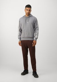Grå hettegenser med frontlomme og sølvstripete ermer, kombinert med mørkebrune joggebukser og svarte sko.