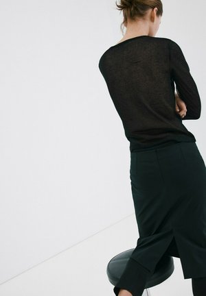 Persona con i capelli legati in piedi con le braccia incrociate, indossando una maglia nera trasparente e una gonna nera, leggermente appoggiata a uno sgabello nero.