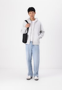 Felpa grigia con zip in tessuto morbido, t-shirt bianca sotto, jeans azzurri chiari, borsa a tracolla nera e sneaker grigie e bianche.