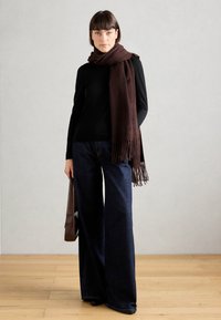 Haut à manches longues noir, jeans larges bleu foncé, et une écharpe à franges marron. Femme tenant un sac marron. Sol en bois, mur neutre.