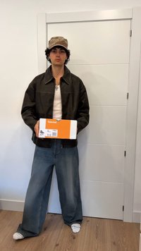 Must must harja, valge särk, laiad teksapüksid ja beež kamuflaažimüts. Hoides valget Timberlandi jalanõude kasti, millel on oranžid detailid.