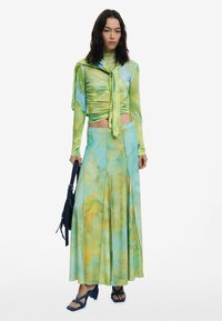 Top a maniche lunghe con nodo frontale in verde e blu, abbinato a una gonna fluida in azzurro chiaro con motivi gialli e verdi, completato da accessori in navy.