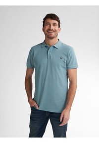 Camisa polo de color azul claro hecha de algodón, con cuello, mangas cortas y un pequeño logo redondo en el lado izquierdo del pecho. Combinada con jeans oscuros.