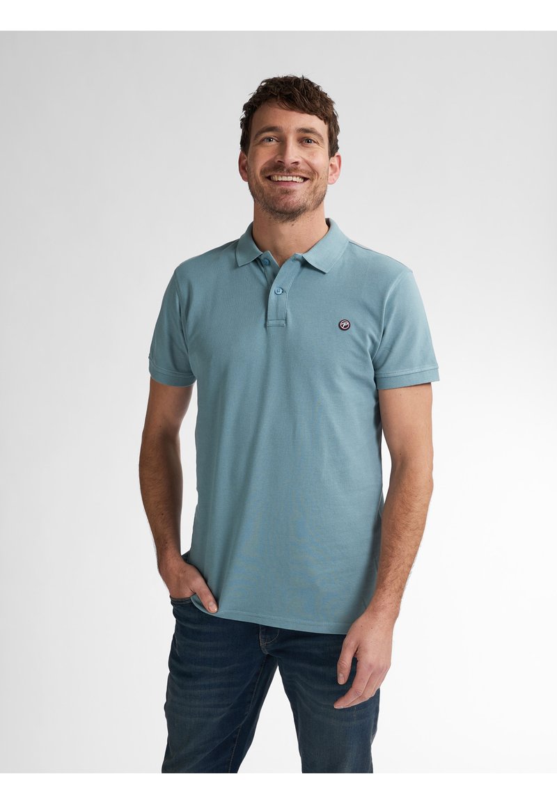 Camisa polo de color azul claro hecha de algodón, con cuello, mangas cortas y un pequeño logo redondo en el lado izquierdo del pecho. Combinada con jeans oscuros.