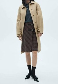 Manteau trench beige sur une chemise sombre, associé à une jupe écossaise marron avec des sangles et des boucles noires, des chaussettes noires et des chaussures à bouts pointus.