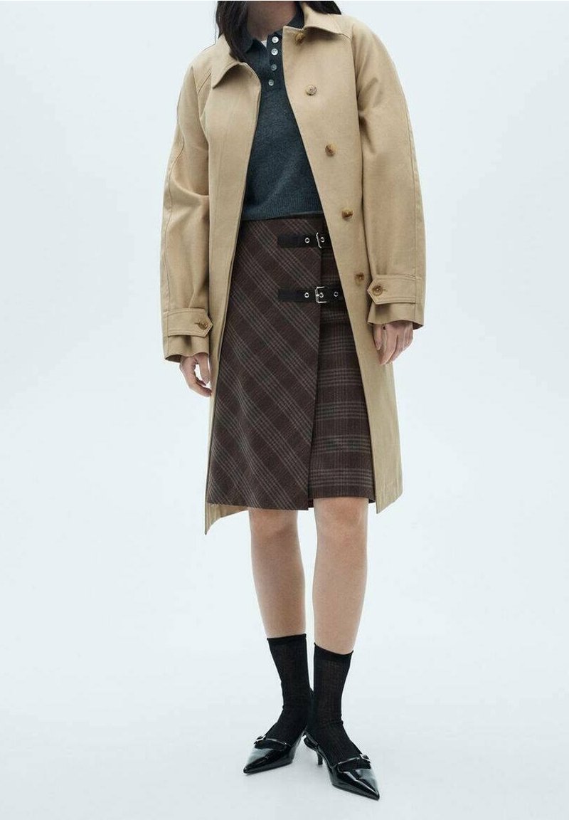 Manteau trench beige sur une chemise sombre, associé à une jupe écossaise marron avec des sangles et des boucles noires, des chaussettes noires et des chaussures à bouts pointus.