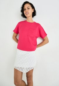 Chemise fuchsia à manches courtes avec une poche poitrine, associée à une jupe mini blanche en dentelle avec un ourlet en forme de scallop. Fond uni.