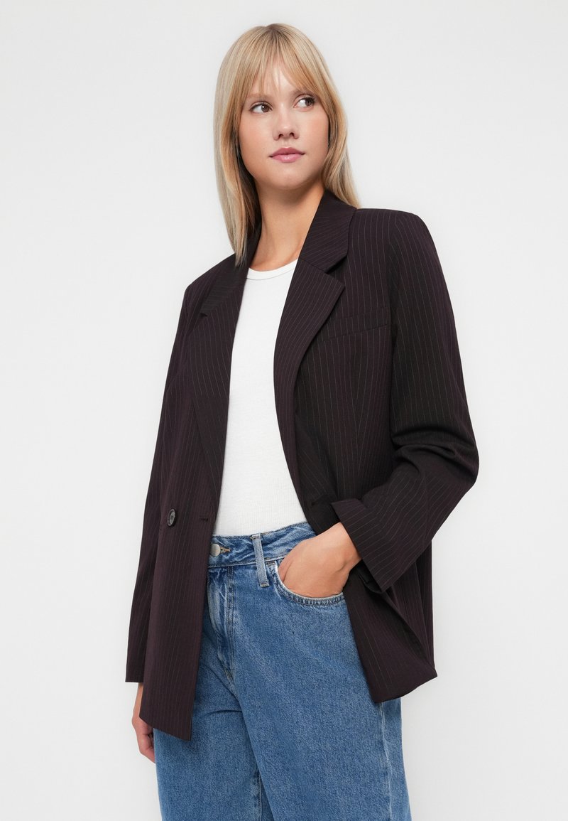 Blazer a righe scure con una vestibilità rilassata, caratterizzato da rever a punta, chiusura con un singolo bottone e due tasche frontali, indossato sopra una maglietta bianca.