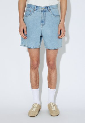 Lichtblauwe denim shorts met vijf zakken, riemlussen en een rafelige zoom. Draagt bij een wit shirt en beige sneakers, gecombineerd met witte sokken.