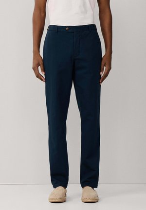 Hombre con pantalones rectos azul oscuro, camiseta blanca y zapatos beige sin cordones, de pie contra un fondo claro y liso.