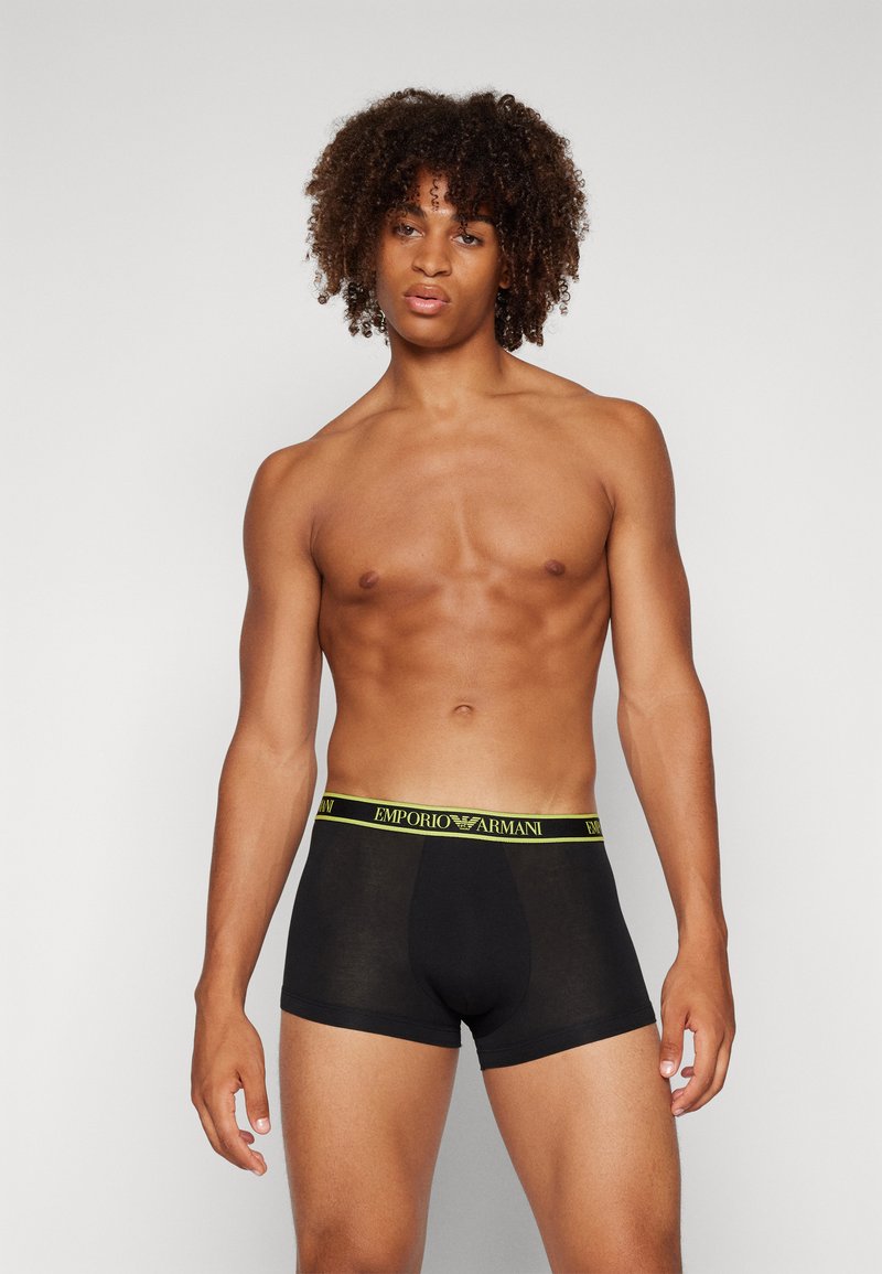 Boxer aderenti neri con elastico giallo che presenta la scritta "EMPORIO ARMANI". Texture liscia, design aderente, lunghezza a metà coscia.