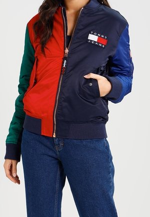 Persoon die een meerkleurige Tommy Jeans bomberjas draagt met rode, marineblauwe, groene en blauwe panelen, gecombineerd met blauwe jeans, hand in de zak.