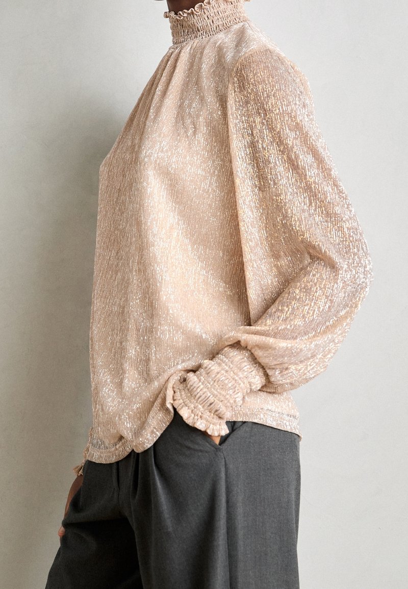 Personne portant un chemisier beige scintillant à manches longues avec un col haut et des poignets à volants, associé à un pantalon ample gris foncé.