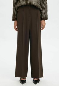 Personne portant un pantalon marron taille haute à jambes larges, associé à des escarpins noirs à bout pointu et un pull marron à motif chevron.