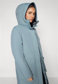 Parka lunga azzurra chiara con cappuccio. Realizzata in tessuto resistente all'acqua, presenta una zip frontale, tasche laterali e una texture liscia.