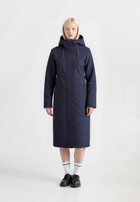 Marineblauwe lange jas met hoge kraag en capuchon, voorzien van ritszakken en een gestructureerde stof voor warmte. Draag met sokken en schoenen.