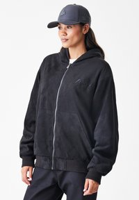 Crna zip-up dukserica od mekog materijala nalik na suede, s kapuljačom, rebrastim manžetama i izvezenim logom na prsima. Uparen s odgovarajućom kapom.