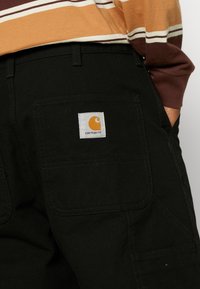 Pantalones cargo negros de tejido duradero, con dos bolsillos traseros y una etiqueta cuadrada de Carhartt con un logo naranja.