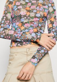 Långärmad, figurnära topp i genomskinligt tyg med ett livligt blommönster i rosa, blått, orange och grönt samt med scalloped kanter.