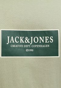 Tejido verde claro con una etiqueta rectangular verde oscuro y texto en blanco. La etiqueta incluye "JACK&JONES", "CREATIVE DEPT. COPENHAGEN" y "©1990."