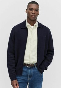 Ung man i en marinblå zip-up cardigan över en krämfärgad skjorta med knappar, blå jeans och ett brunt flätat bälte, stående med ena handen i fickan.