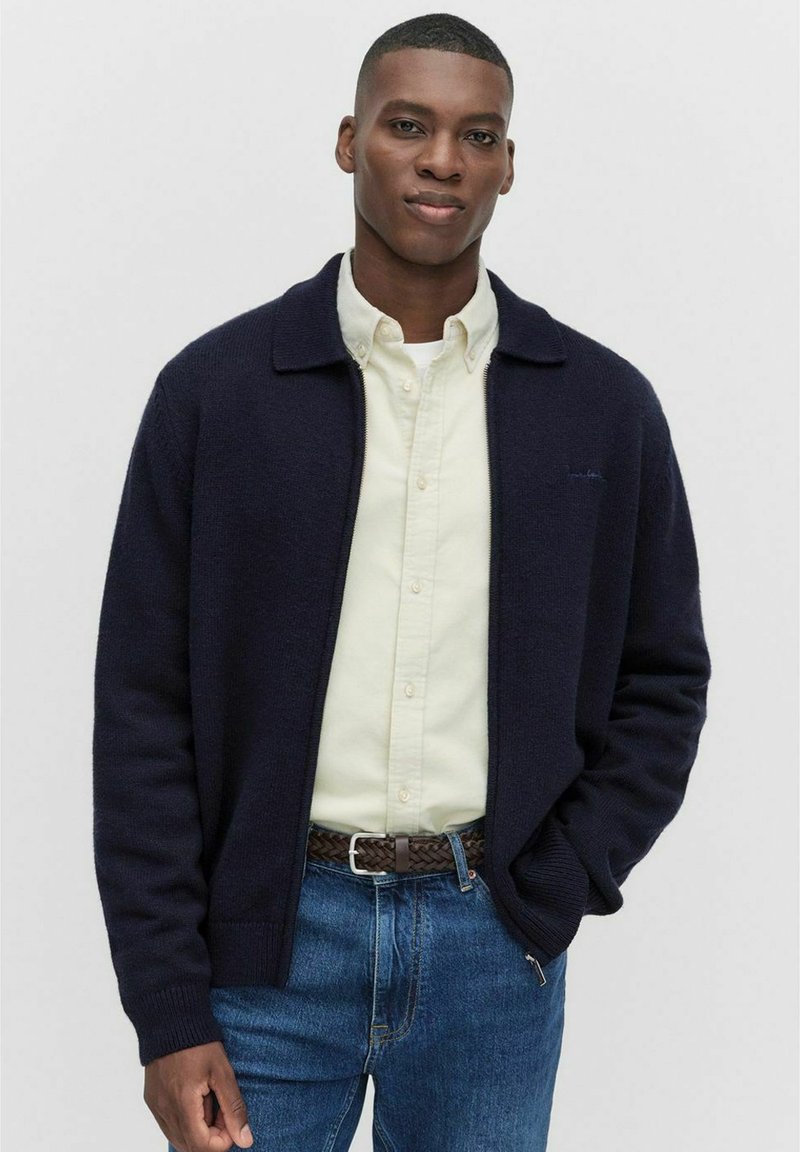 Ung man i en marinblå zip-up cardigan över en krämfärgad skjorta med knappar, blå jeans och ett brunt flätat bälte, stående med ena handen i fickan.