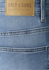 Jasnoniebieskie dżinsy z denimu z teksturowaną skórzaną metką z wytłoczonym napisem „ONLY & SONS”. Widoczne przeszycia podkreślają szczegóły projektu.