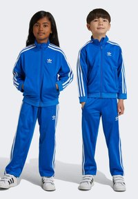Blå polyester träningsoverall med vita ränder på ärmar och byxor, med Adidas-logotyp på bröstet och benet. Avslappnad passform, framfickor.