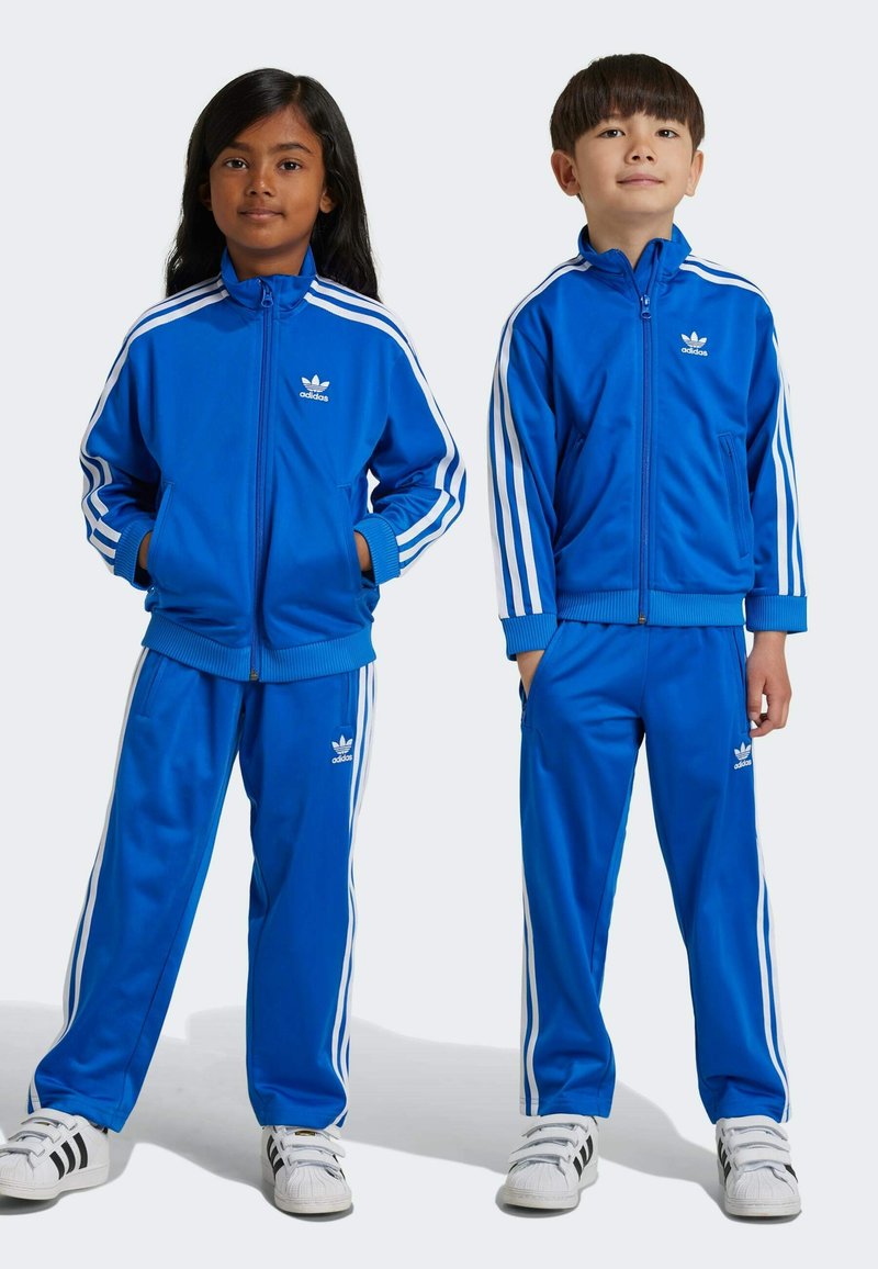 Blå polyester träningsoverall med vita ränder på ärmar och byxor, med Adidas-logotyp på bröstet och benet. Avslappnad passform, framfickor.