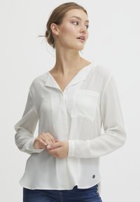 Blusa bianca a maniche lunghe in tessuto leggero con tasca sul petto sinistro e orlo curvo. Presenta una chiusura con bottoni e una morbida consistenza.
