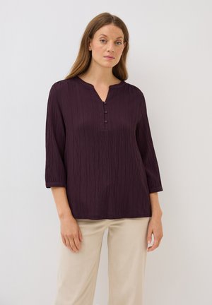 Chemise à manches longues violet foncé avec un tissu texturé, col rabattu et trois boutons au niveau de la poitrine. Portée avec un pantalon beige clair.