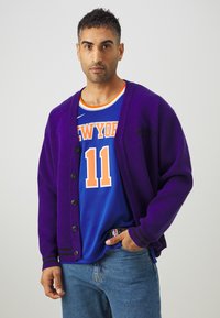 Lilla cardigan med knapper, prydet med sorte striber. Båret over en blå basketballtrøje med orange bogstaver og nummer.