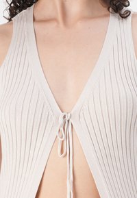 Haal beige ribbattu tank top, jossa on syvä V-kaula-aukko, ohuet hihnat ja etutaskun suljin. Pehmeä materiaali ja avonainen selkämallitus.