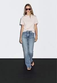 Helles rosa T-Shirt, lockere blaue Jeans und schwarze Flip-Flops. Das Model trägt eine Sonnenbrille und steht vor einem schlichten weißen Hintergrund.